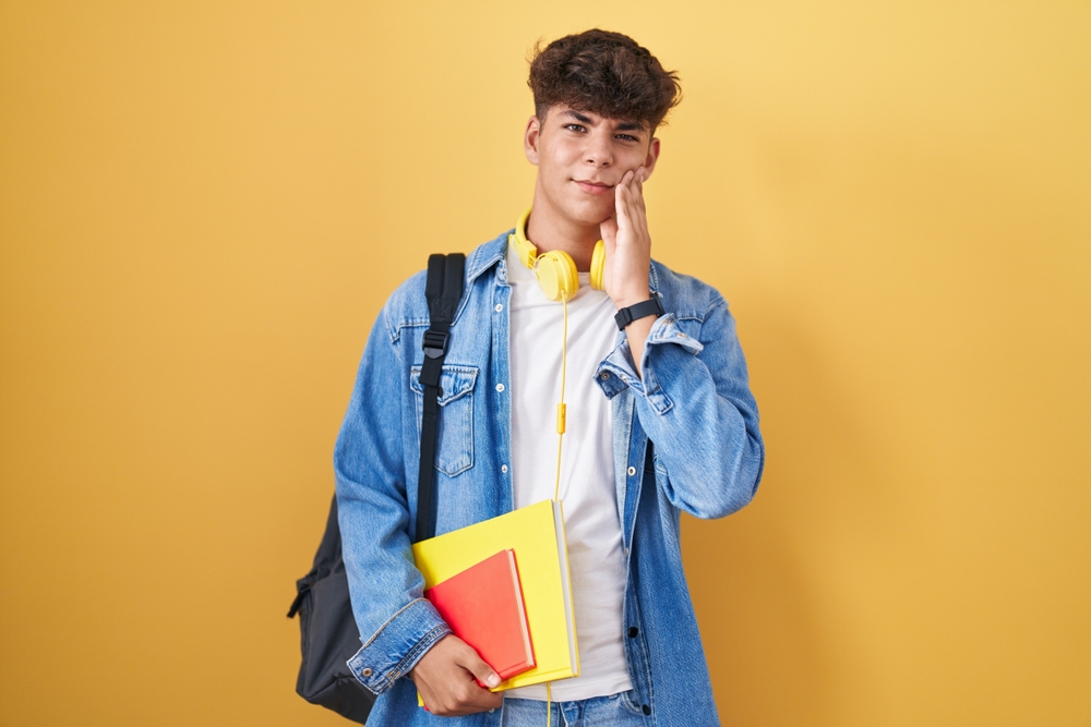 Hispanic,Teenager,Wearing,Student,Backpack,And,Holding,Books,Touching,Mouth